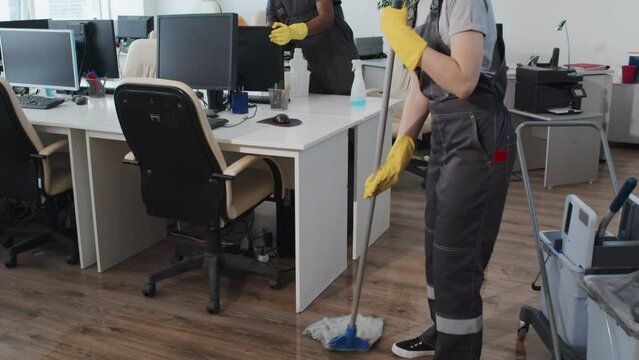 Selective focus slowmo of Caucasian woman in uniform washing floor using mop, her Black male co-worker cleaning computer monitor and desk