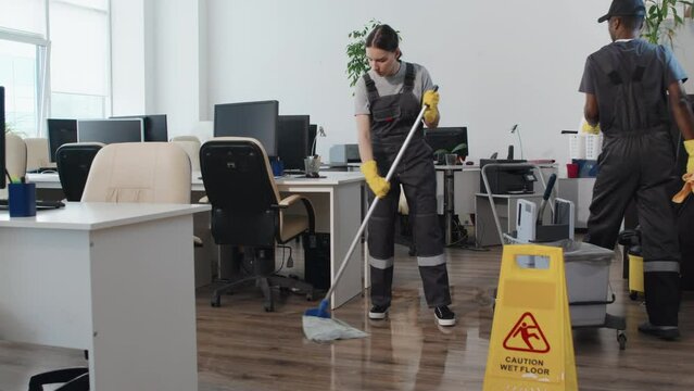 Slow motion long shot of young Caucasian professional cleaner mopping floor, her Black co-worker bringing detergents to wipe computer monitors