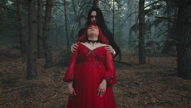 Portrait goth man vampire king in black tailcoat hugs hypnotized redhead woman queen. fantasy sleep sexy dream. Romantic love couple two evil face. Red dress. Mystery night fog forest dark tree 4k
