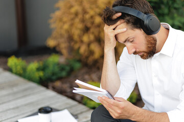 Obraz premium Thoughtful bearded man in headphones checking notebook, thinking in park.