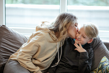 Happy mother and school aged son sitting on sofa near window. Woman kiss to cheek little boy,...
