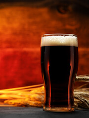 A glass of dark beer on a wooden table with ears of wheat. Back light