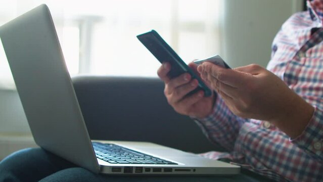 man pressed credit card code to pay online via smartphone while sitting on sofa at home with laptop in room, online shopping and using credit cards concept close-up.
