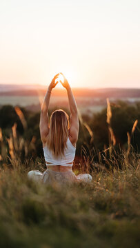 Back View on Woman Sitting in Meditation Yoga Pose and Catching Sun by Hands at Sunset Outdoors