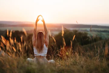 Fotobehang Back View on Woman Sitting in Meditation Yoga Pose and Catching Sun by Hands at Sunset Outdoors © Romvy