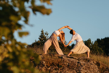 Young Adult Couple Making Beautiful Pose, Man and Woman Practicing Yoga Outdoors on the Rocky Hill at Sunset