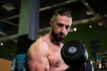 Fototapeta na wymiar A handsome muscular man with a beard looks into the camera and does a bicep exercise with a dumbbell. Sports, healthy lifestyle