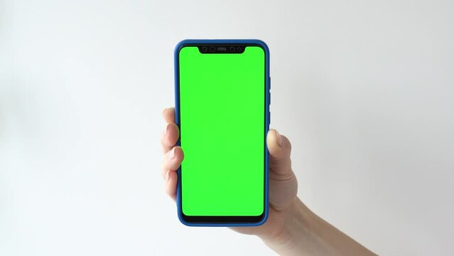 Hand of young woman holding a smartphone with vertical green chroma key screen on gray backdrop. 4k template. Girl hands keeps cell phone with green mockup screen background in front of the camera