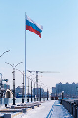 The flag of the Russian Federation on a high flagpole in winter on a snow-covered embankment. Frosty morning. Lanterns and benches. The crane behind the fence at the construction site