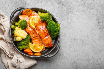 Healthy baked fish salmon steaks, broccoli, cauliflower, carrot in black cast iron casserole bowl on grey stone background. Cooking a delicious low carb dinner, healthy nutrition, space for text - 569998836