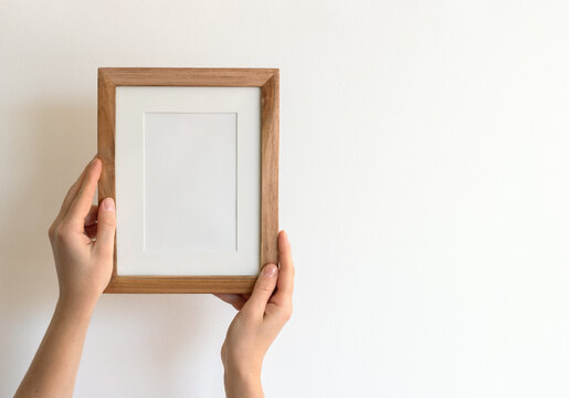 Woman's hands holding a wooden stylish blank frame against a white wall. Frame mock up, copy space for your text.	
