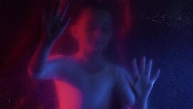 Trapped child. Defocused silhouette. Freedom isolation. Red blue neon light scared small girl hands touching creased texture transparent polyethylene film on dark out of focus.
