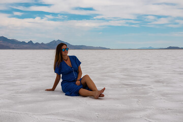 Woman in blue dress sitting on Bonneville Salt Flats near Wendover, Western Utah, USA, America. Salt pans and barren landscape near Salt Lake City. Panoramic view of Silver Island Mountain peaks