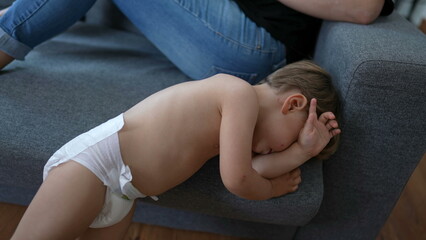 One tired little baby toddler wearing diapers rubbing face on couch - obrazy, fototapety, plakaty