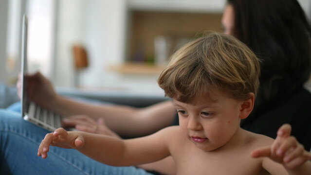 Toddler Boy Plays With Hair Tier While Mother Works From Home In Front Of Laptop At Home Sofa In Background. Little Boy Stretching Elastic Band. Parent Multi Tasking