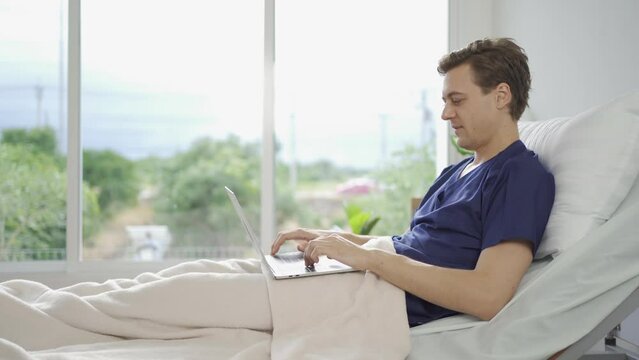 Portrait of happy caucasian sick patient using a computer notebook laptop device to work on business on bed in hospital in medical and healthcare treatment at nursing home or clinic. People lifestyle