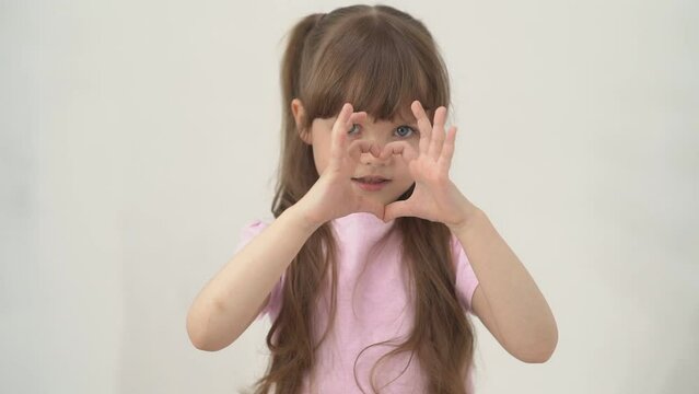 Portrait of a funny little girl with brown hair and a toothless smile, holding her heart-shaped hands at her mouth with her baby teeth falling out. The concept of healthy teeth in childhood. Copy