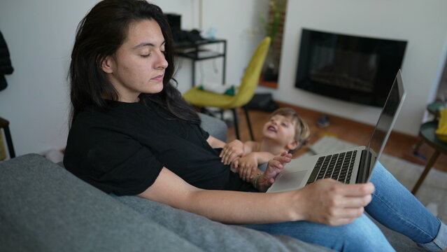 Child Interrupting Mother Working From Home In Front Of Laptop. Parent Multi Tasking Using Computer Sitting On Couch. Parenting And Multi Task