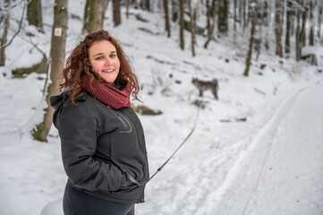 Young woman with brown curly hair and warm clothing is smiling and looking at camera, walks her gray colored akita inu dog in the forest during winter with lots of snow