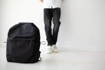 The guy with a bagpack on his shoulders is poaing  on a white background. The teenager  is wearing a white T-shirt and black jeans