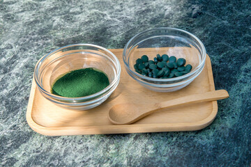 spirulina powder and tablets in glass bowls on a wooden tray with a wooden spoon on a green marble background.