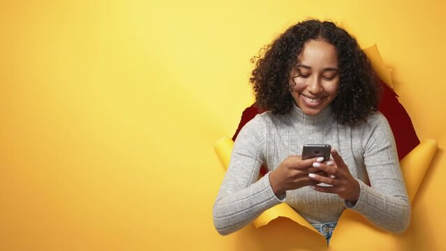 Mobile app. Online chat. Social media communication. Happy smiling woman using phone texting message in orange breakthrough hole torn paper wall copy space background.