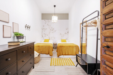 Fototapeta premium Bedroom with single beds separated by a wooden stool with mustard colored blankets and a black dresser with drawers
