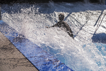 A surfer in the water giving it a go in a wave pool with big splashes in the turns and sunlit drops everywhere