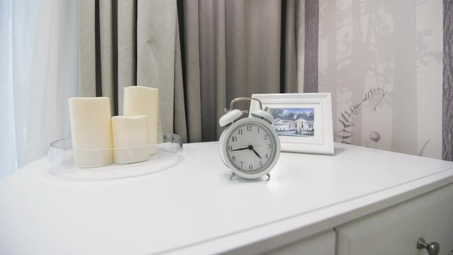 Alarm clock on white chest of drawers near candles and photo frame in room made in white and greyish shades. Minimalist interior in luxury house