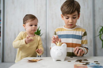 Siblings boy and girl small caucasian child brother and sister play at home with piggy bank saving...
