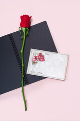 Red rose and blank black paper book on pink background with blank greeting card. Copy space.