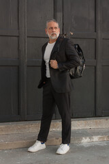 Attractive gray-haired man dressed in black suit walking downtown with black backpack