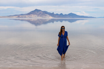 Woman in blue dress walking into lake Bonneville Salt Flats, Wendover, Western Utah, USA, America. Beautiful summits of Silver Island Mountain range reflecting in water surface west of Great Salt Lake