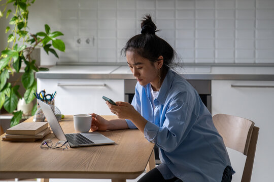 Young Concerned Self-employed Asian Woman Sits At Home Office Checking Phone Reading Last News And Drinking Coffee Before Starting Remote Work Day. Female Freelancer Using Mobile Device At Workplace