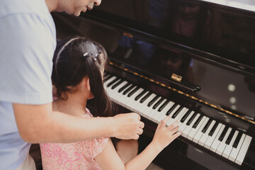 Fototapeta premium Cute and focused young little Asian girl practicing songs and melody with father and tutor while pressing white and black key and creating new music on piano as hobby and dedication at home