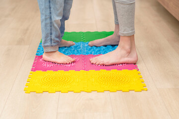 Health concept. Two boys stand barefoot on a colorful orthopedic massage mat in a home interior. Close-up of children's legs.