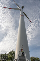 female engineer with brown curly hair, yellow helmet and digital tablet standing in front of the entrance of a wind turbine, low angle view