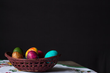 Colored Easter eggs in a wooden basket on a white tablecloth at black background. Copy space.