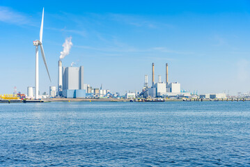 Naklejka premium Coal fired power plants with high smokestacks on a harbour. A tall wind turbine is in foreground.