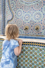 Toddler watches water run from brass faucets in a Moroccan fountain; intricate ceramic tile mosaic