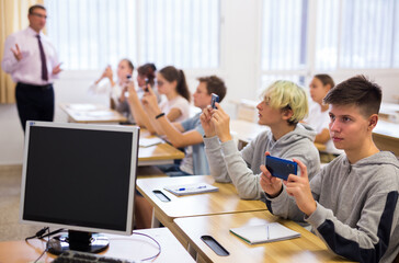 Plakat Group of modern teenagers sitting with mobile phones on lesson in classroom