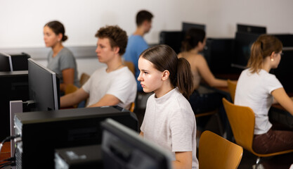 Focused teenager students sitting at library desk in computer room with pc, preparing for exam. Focus on young girl