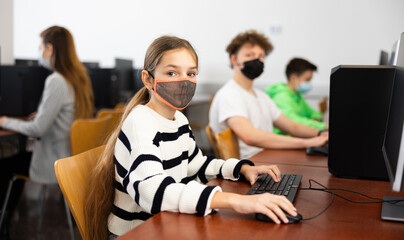 Group of young girls and boys in face masks sitting in computer classroom of library and exercising.