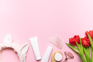 Face care concept. Asian Gua Sha massage tools, cream, mask in white plastic tubes, hair band and beautiful red tulips on light pink background with copy space for your design.