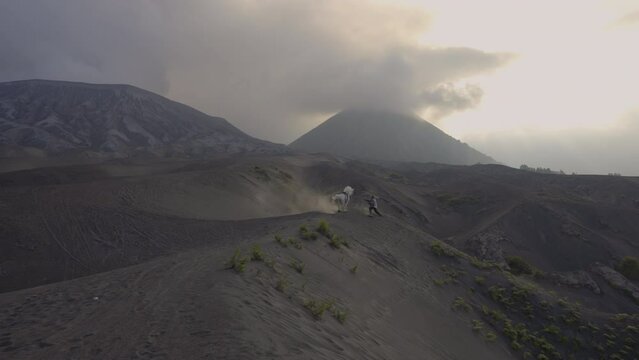 Man Controlling Bucking Horse On Desert Mountain Under Clouds During Sunset - Java, Indonesia