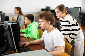 Teenage boy sitting at table and helping young girl with PC problem in computer class.