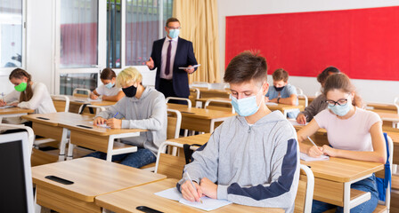 Focused teenage students in protective face masks studying in classroom with teacher, writing lectures in workbooks. Necessary precautions in coronavirus pandemic