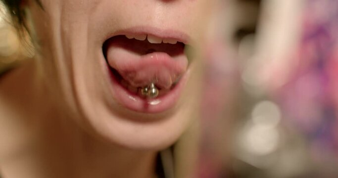 Woman shows tongue piercing into camera phone, smartphone bokeh. Two earrings, she moves her tongue, covers her mouth, smiles.