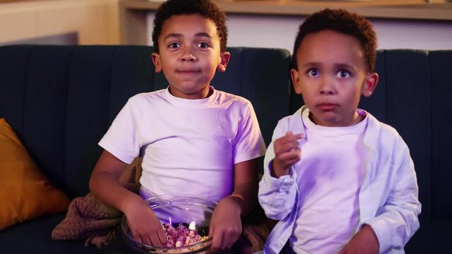 Camera view of cute little african american boys brothers eating popcorn sitting on sofa under the blanket watching movie film show television at late night at home together Family time