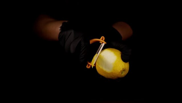 Person in black gloves peels yellow zest from fresh lemon fruit with sharp peeler on deep black background extreme closeup